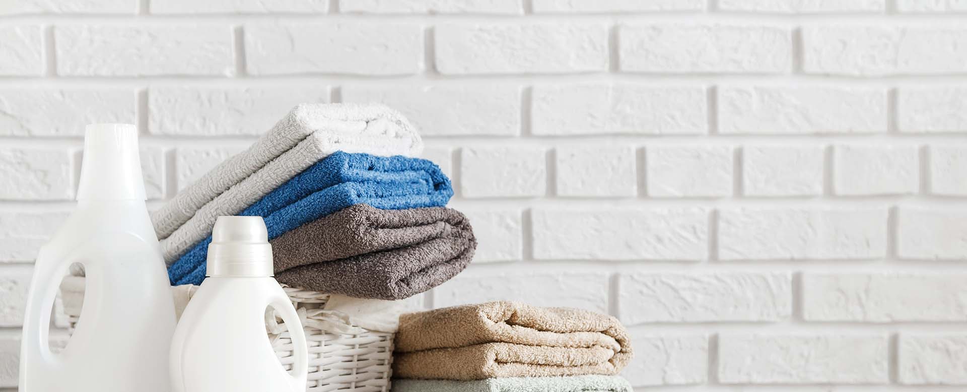 Laundry concept. Bottles with detergent gel and lush towels in in basket on wooden table with white brick wall on background.